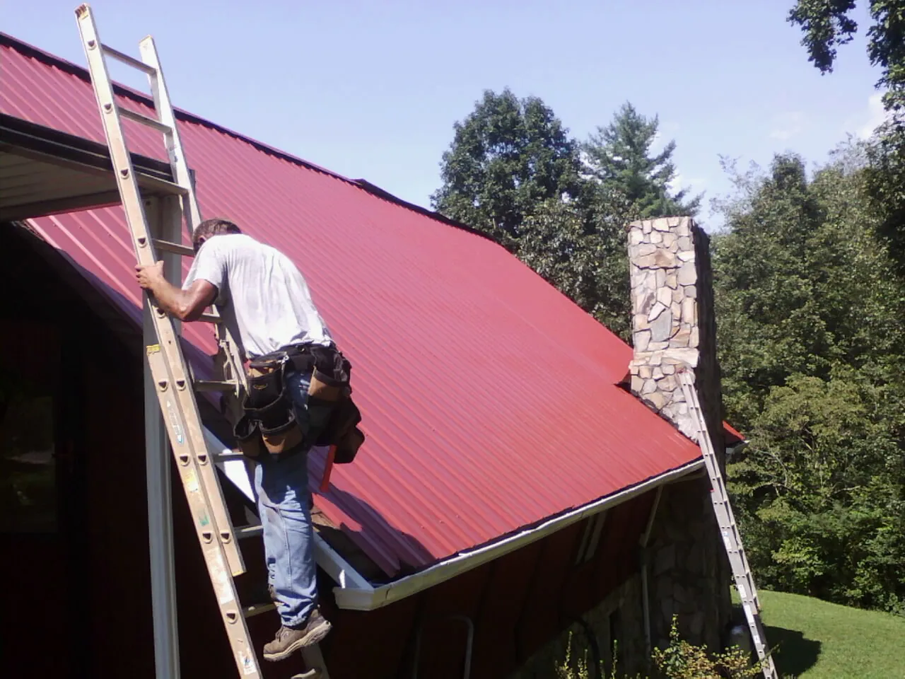 Roofer on a ladder beside a red metal roof and stone chimney — Soffit & Fascia Repair by Roofing Brothers in Cumberland Foreside