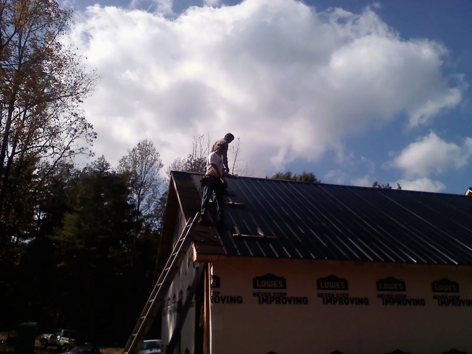 Roofer fastening dark metal panels high against a cloudy sky, from a Roof Inspection job in Cumberland Foreside